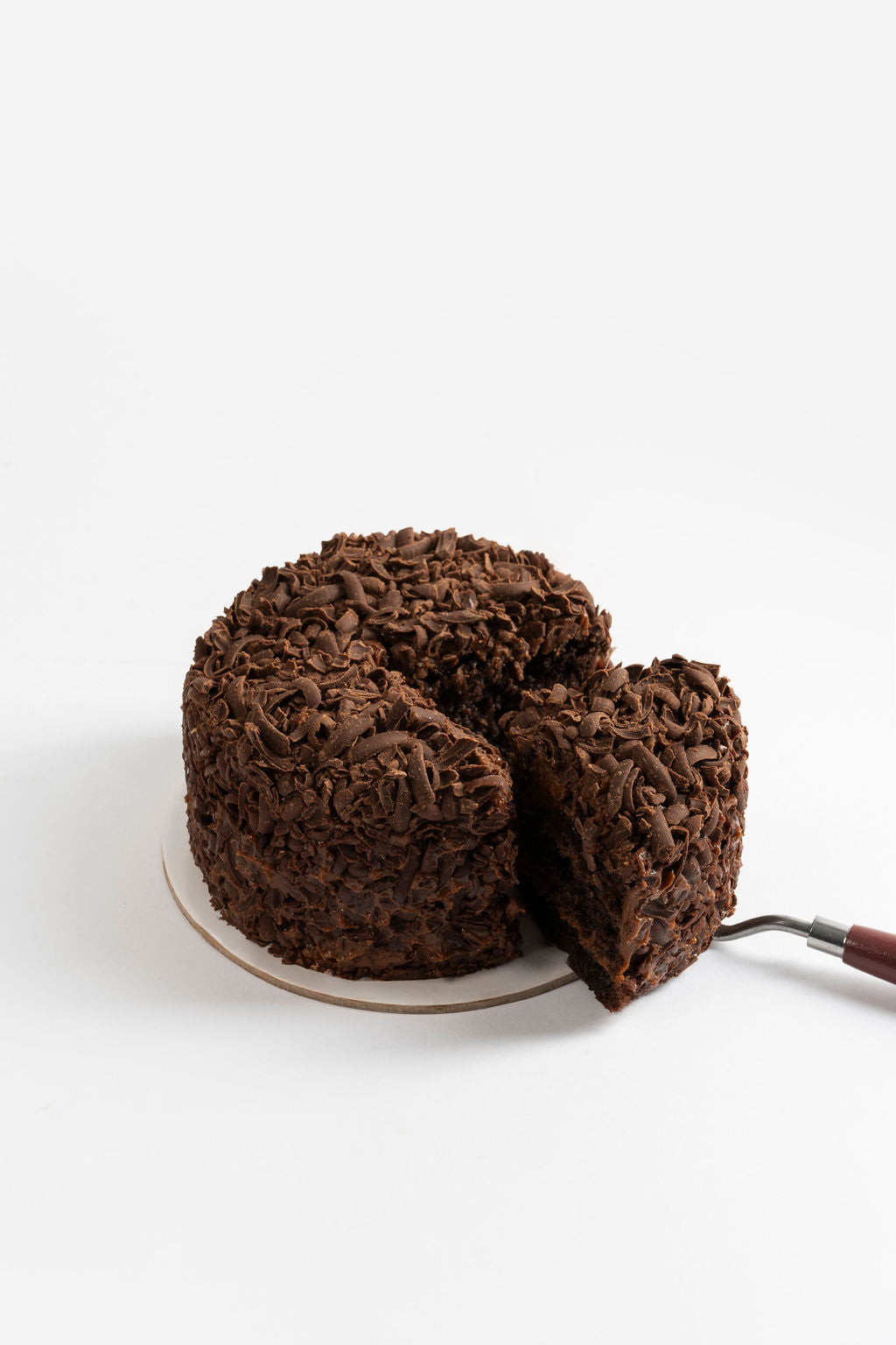 Torta de chocolate con forma circular, cubierto completamente de virutas de chocolate oscuro. Una porción ha sido cortada y está siendo servida, mostrando el interior esponjoso y húmedo